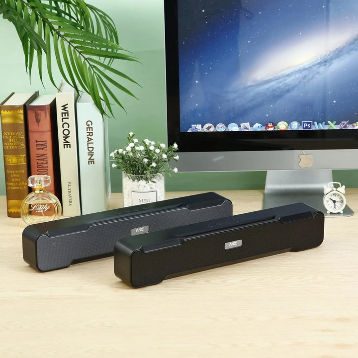 Two black computer speakers on a desk with a monitor, books, and a plant in the background.